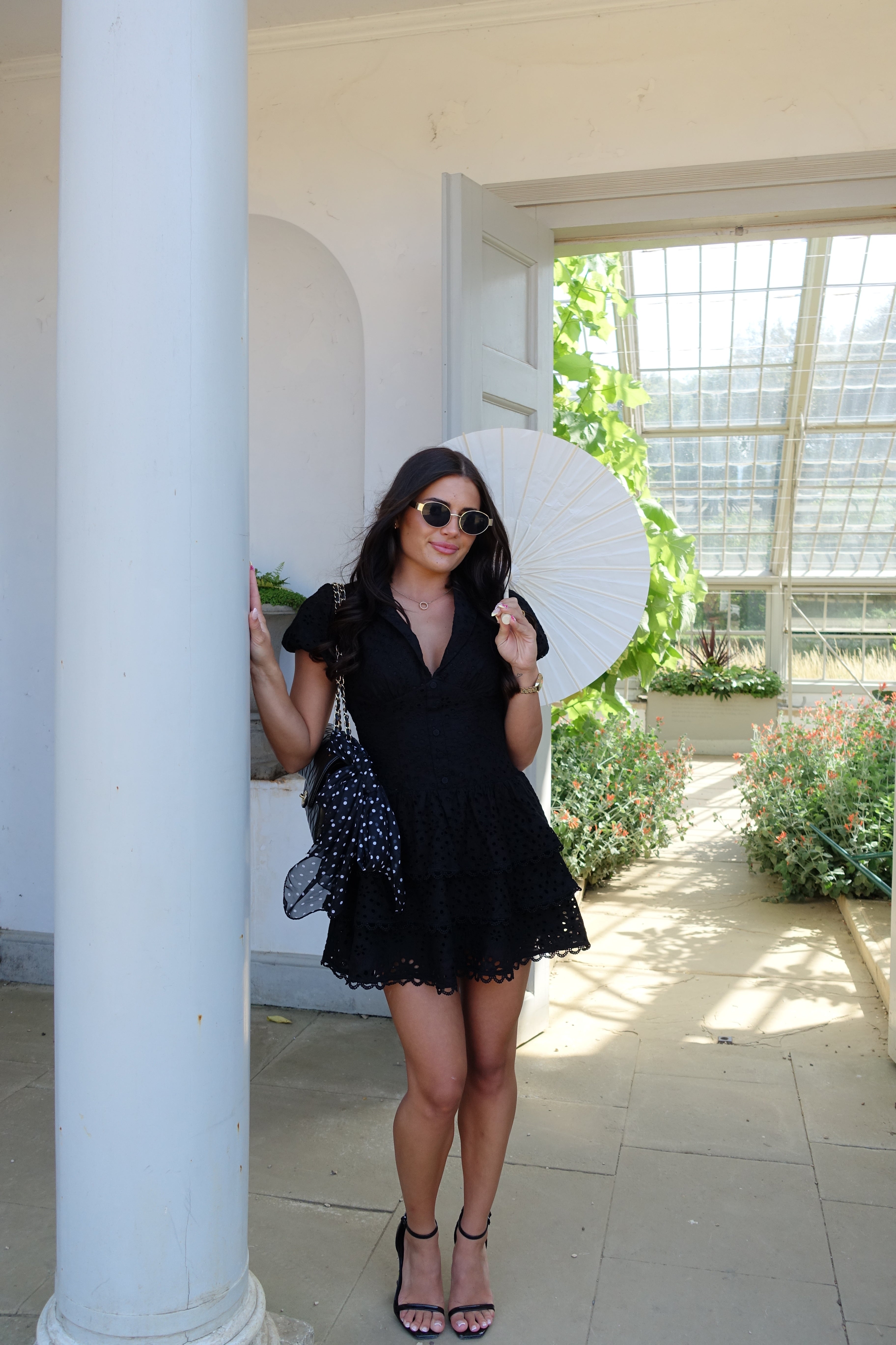 Woman in a black dress and white hat sitting on a white railing with greenery in the background